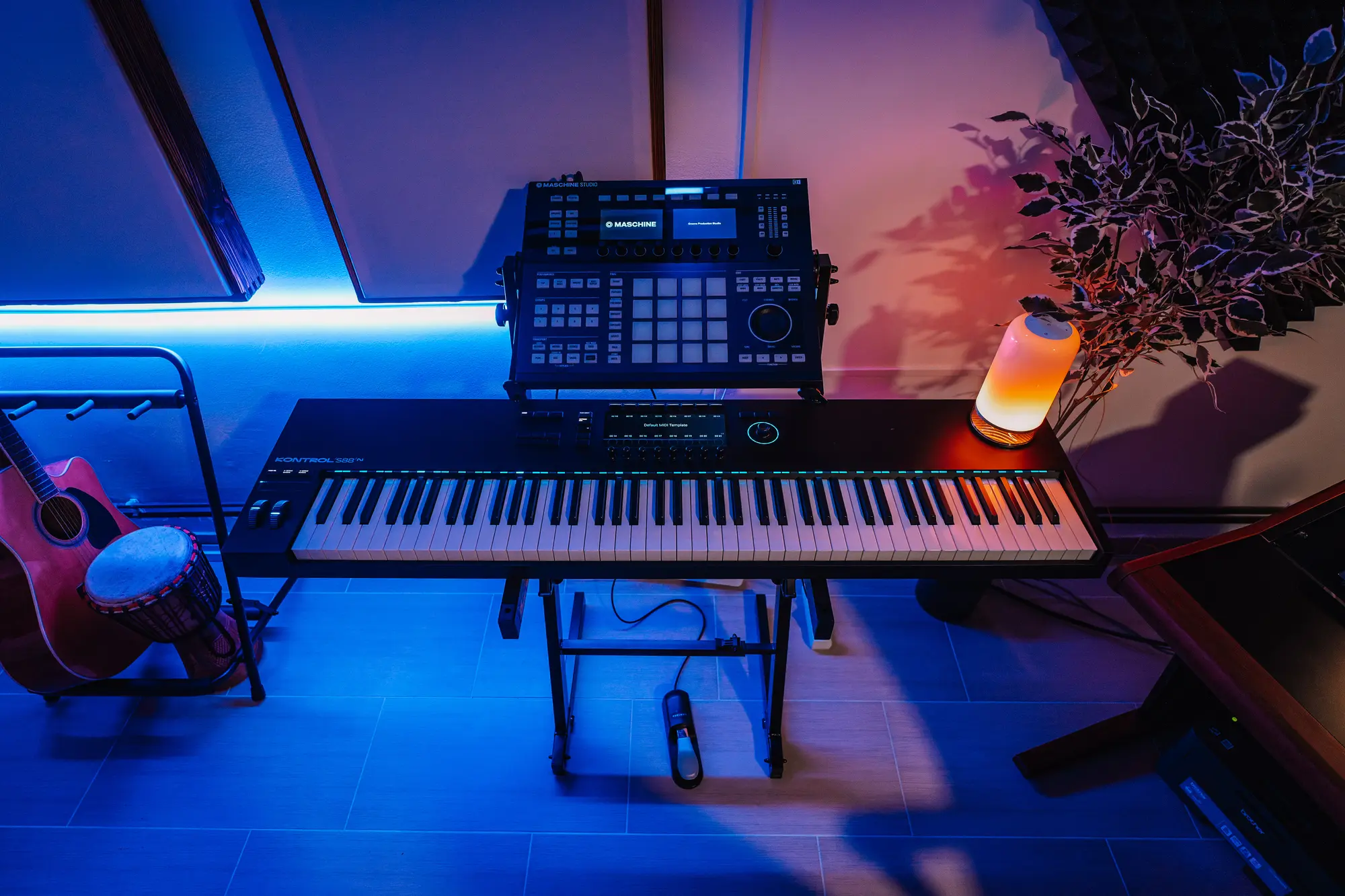 Native Instruments Kontrol S88 with Maschine Studio on a stand, an acoustic guitar and a djembe beside it, under blue and amber light